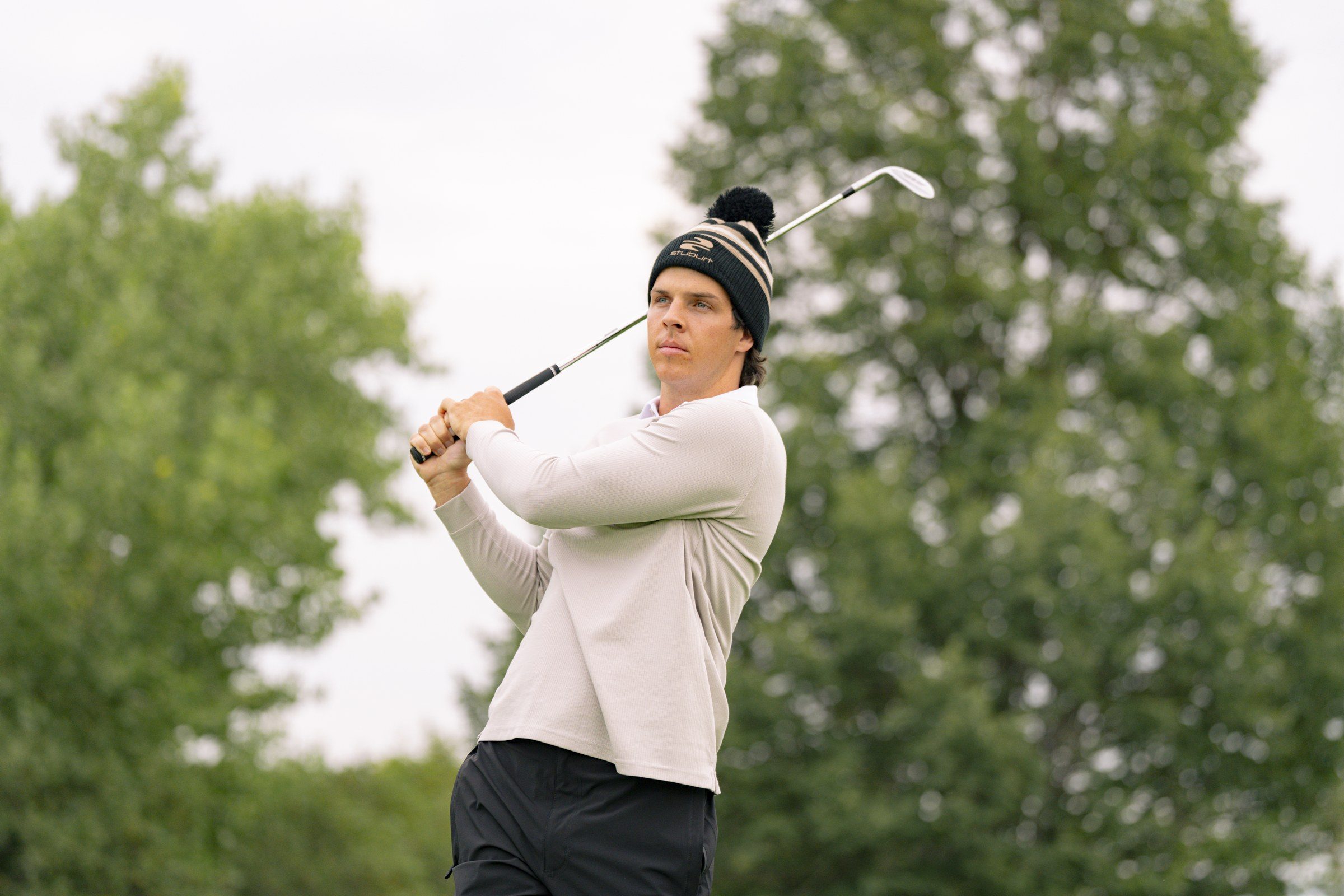Golfer in a Stuburt bobble hat and cream long-sleeve polo following through on a swing